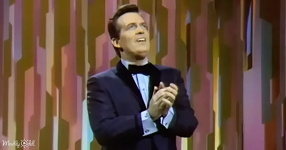 A man in a tuxedo claps, wearing a bow tie, against a colorful striped background.