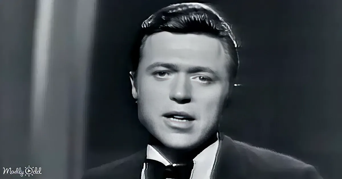 A man in a suit and bow tie, with styled hair, appears to be speaking or singing.