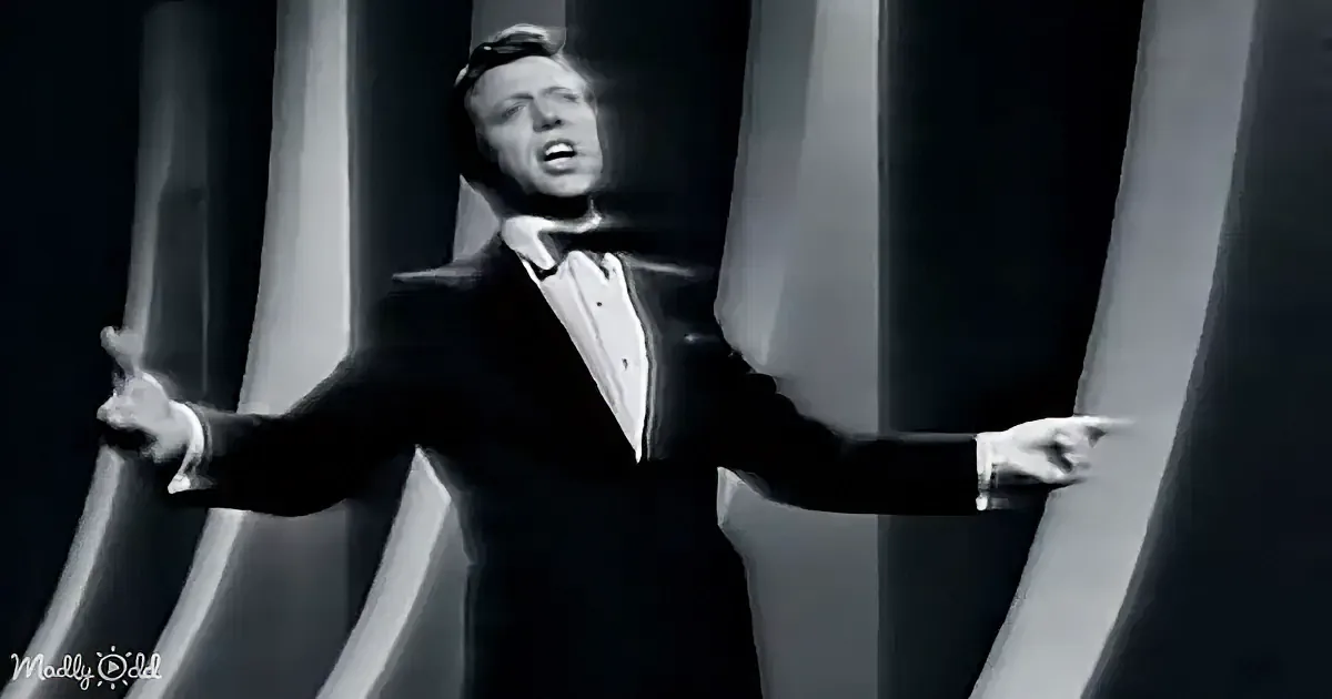 A man in a tuxedo is dramatically gesturing with his arms outstretched, surrounded by large columns.