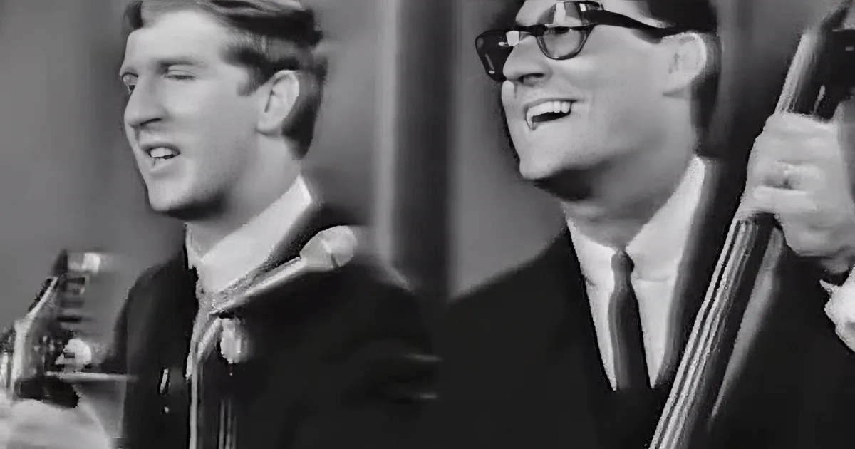 A man and woman are singing together, the man in a suit and tie, the woman with dark hair and bangs.
