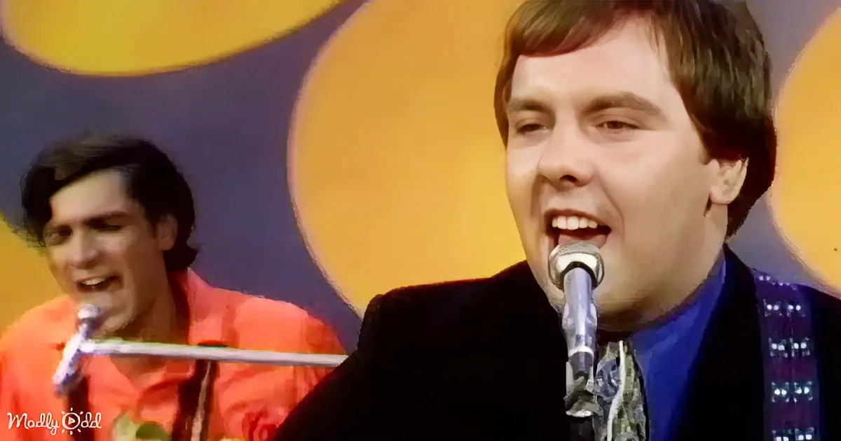 A man in a blue suit sings into a microphone, with another person in a red shirt in the background.