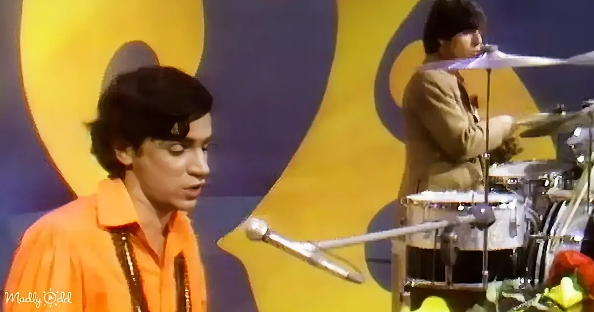 A person in an orange shirt sings into a microphone, with a drummer in a brown suit in the background.