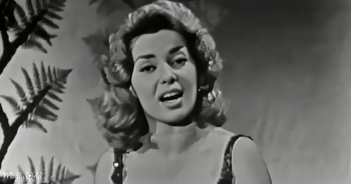 A woman with curly hair, wearing a sleeveless top, appears to be singing or speaking in a black-and-white scene.