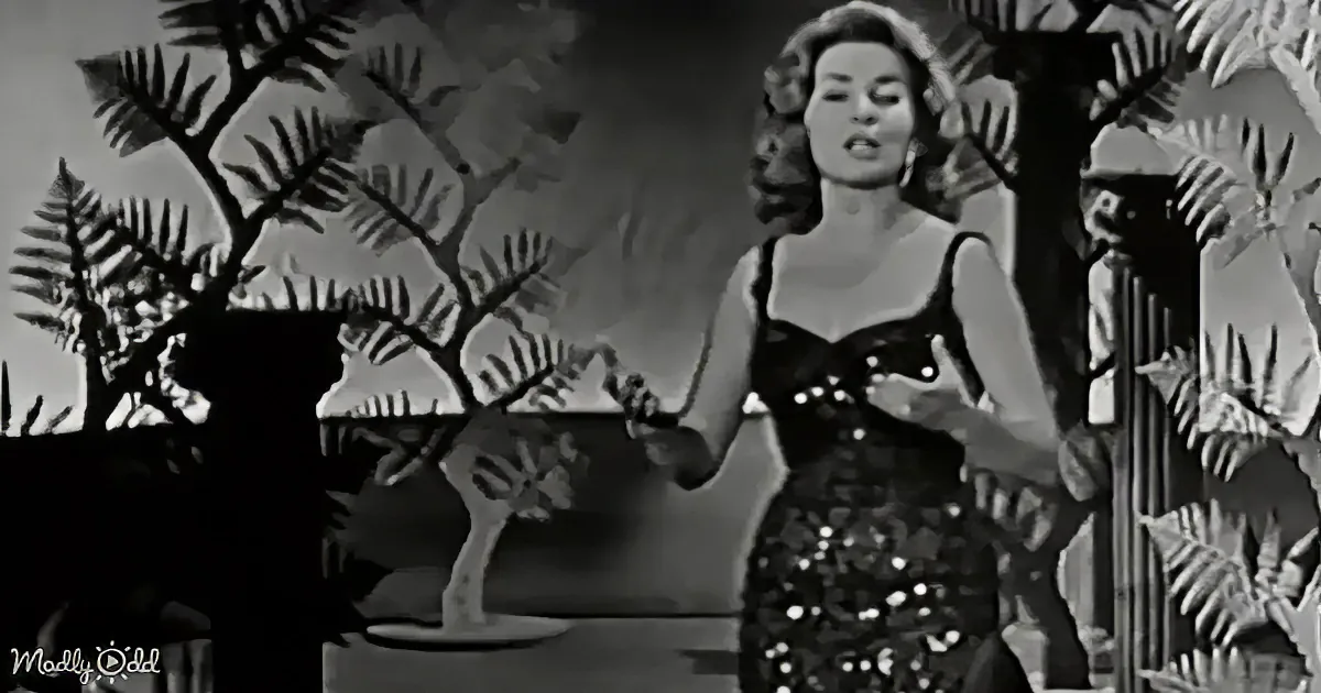 A woman in a sequined dress stands in front of a tropical backdrop, gesturing with her hands.