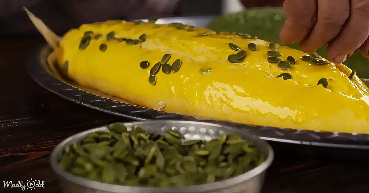 A hand sprinkles pumpkin seeds on a yellow, elongated dessert on a black plate.