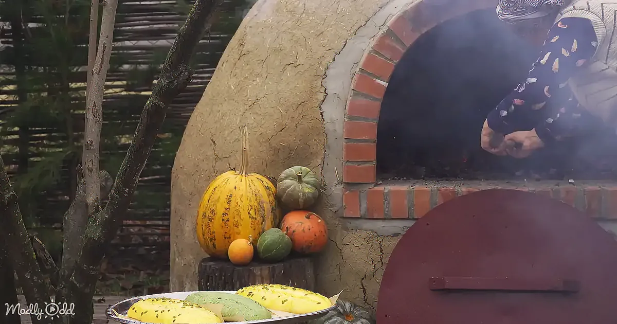 A person tends to a brick oven, surrounded by pumpkins and colorful breads on a table.