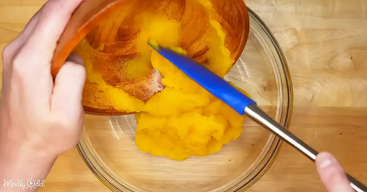 A hand holds a pumpkin while a knife scoops out its flesh into a glass bowl.