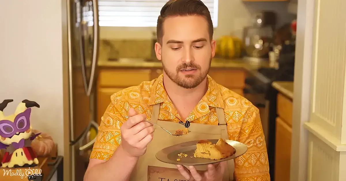 A man in a yellow shirt and apron holds a plate with a piece of cake, eating with a fork.
