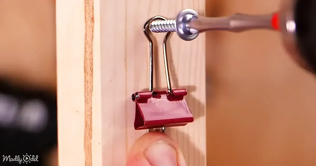 A red binder clip is being held by a hand, with a tool about to screw it onto a wooden surface.