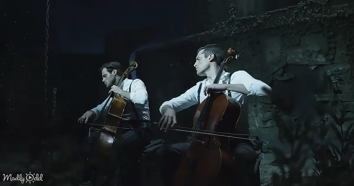 Two men in white shirts play cellos in a dimly lit, stone-walled room.