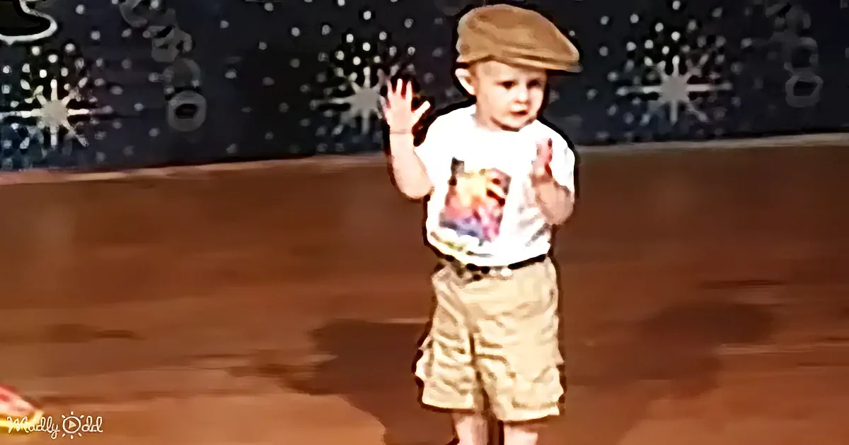 A young child wearing a brown cap and beige shorts stands on a stage, waving with one hand.