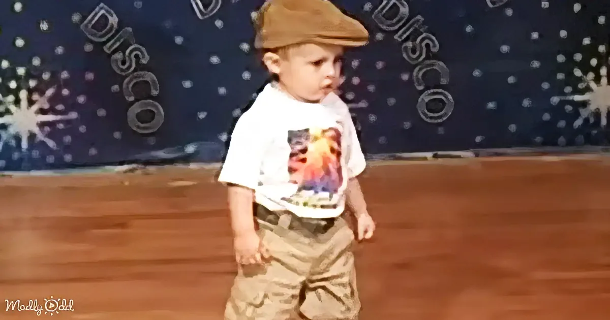 A young child wearing a brown cap and a white shirt with a graphic design stands on a wooden floor in front of a...