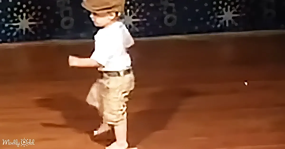 A young child in a white shirt and khaki shorts stands on a wooden floor, wearing a hat and sunglasses.