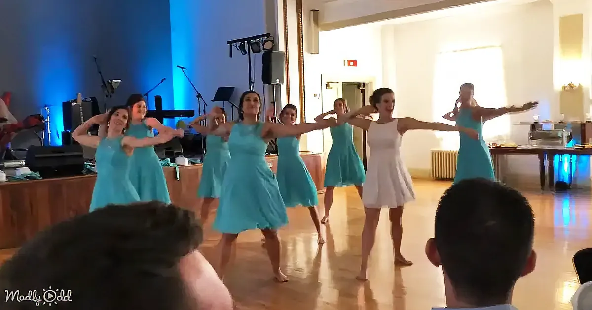 A group of women in blue dresses perform a synchronized dance in a brightly lit room with a stage and audience in the...