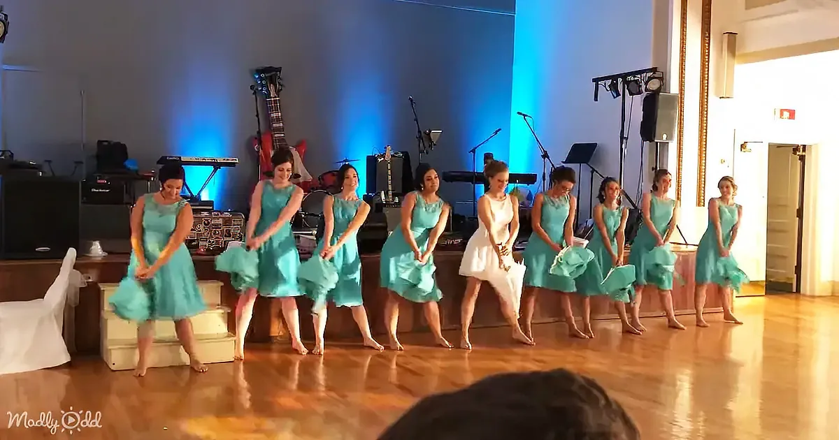 A group of women in teal dresses performs a synchronized dance on a wooden floor.