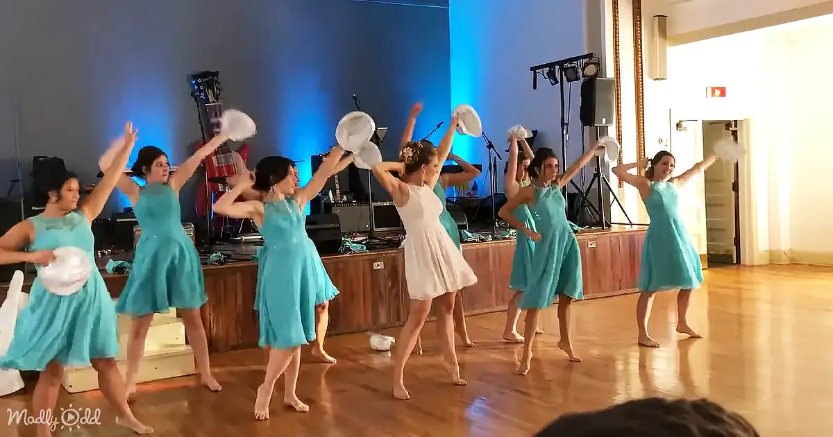 A group of women in teal dresses performs a synchronized dance with white hats, in a room with a wooden floor and stage...