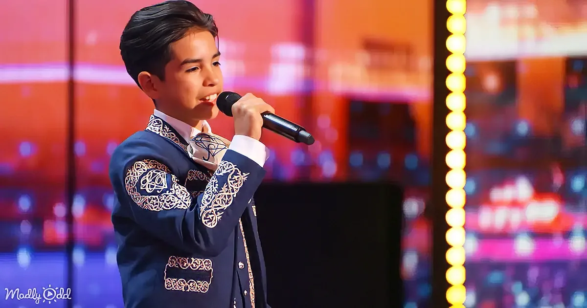 A young boy in a blue embroidered suit sings into a microphone on a stage with colorful lights.