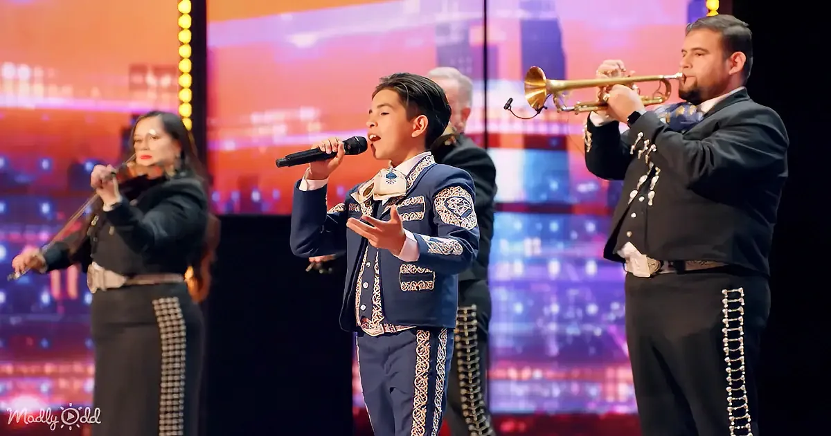 A young boy in a blue mariachi outfit sings into a microphone on stage, accompanied by musicians playing instruments.