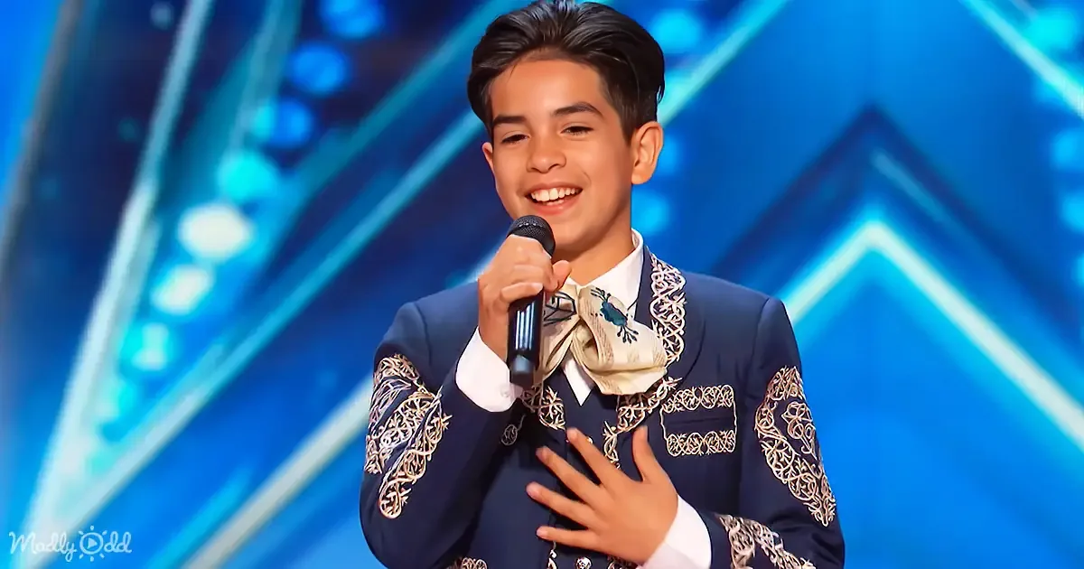 A young boy in a blue suit with intricate embroidery smiles while holding a microphone on stage.