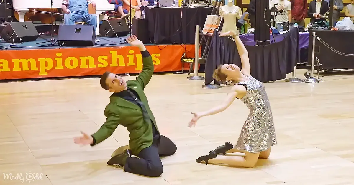 A man and woman in formal attire kneel on a dance floor, arms raised, during a dance competition.