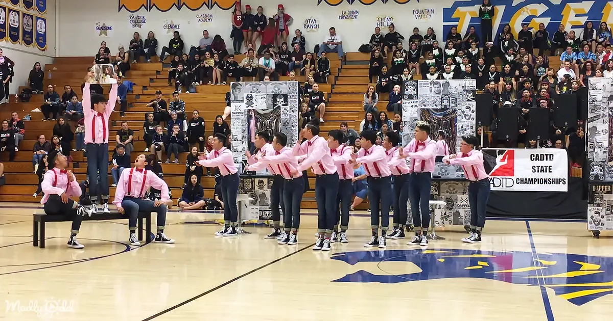 A group of dancers in pink shirts and red suspenders perform on a basketball court, with spectators in the background.