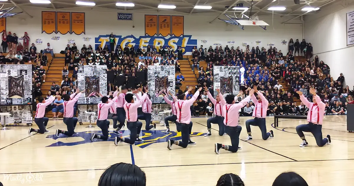 A group of people in pink shirts and red suspenders kneel on a basketball court, raising their hands in unison.