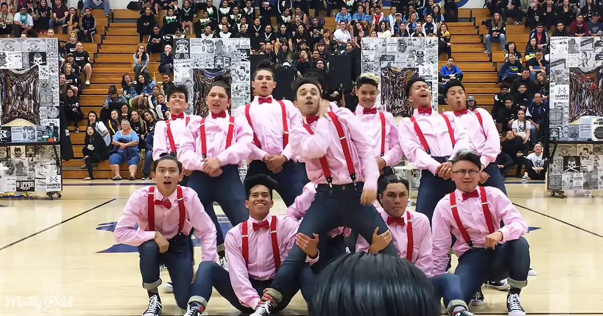 A group of young men in pink shirts and red suspenders perform a synchronized dance on a gymnasium floor.