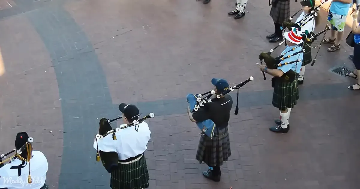 A group of bagpipers in kilts and traditional attire plays on a brick-paved street.