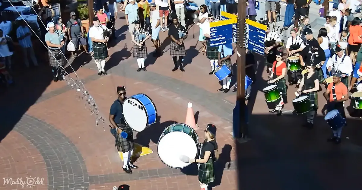 A bagpiper in a kilt plays on a brick plaza, surrounded by people sitting and watching.