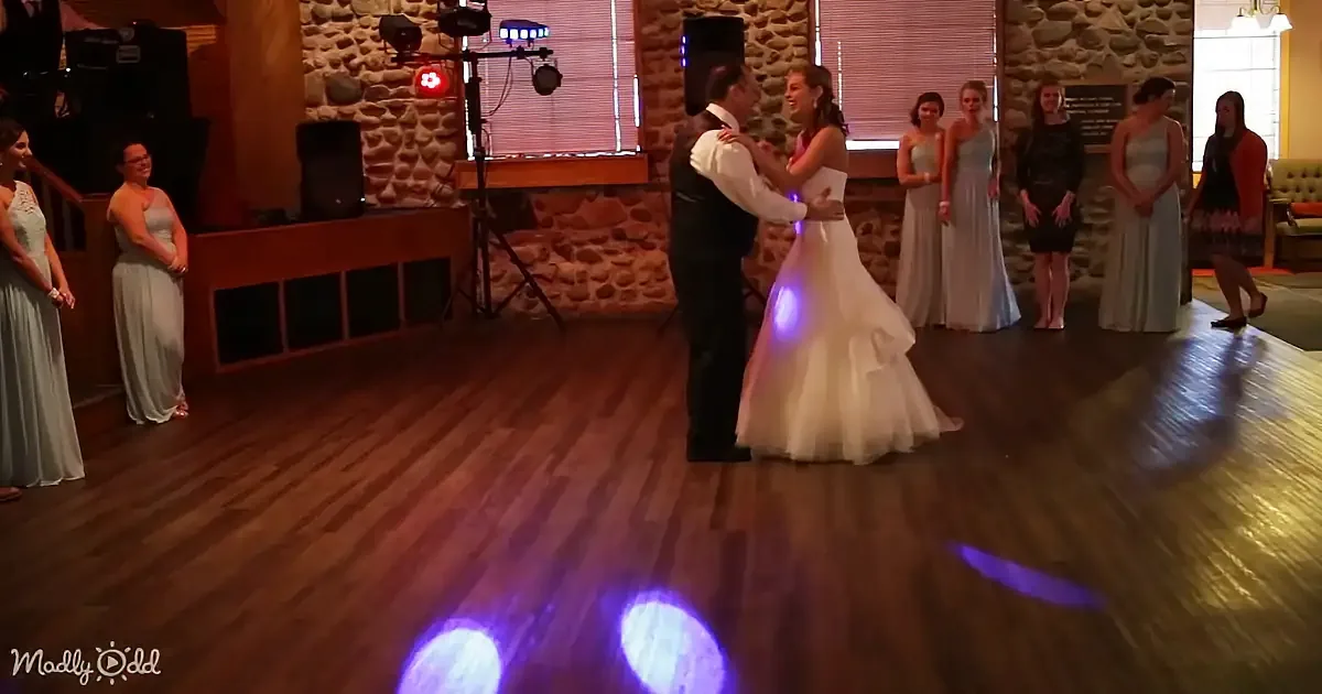 A bride and groom share a dance on a wooden floor, surrounded by guests in formal attire.