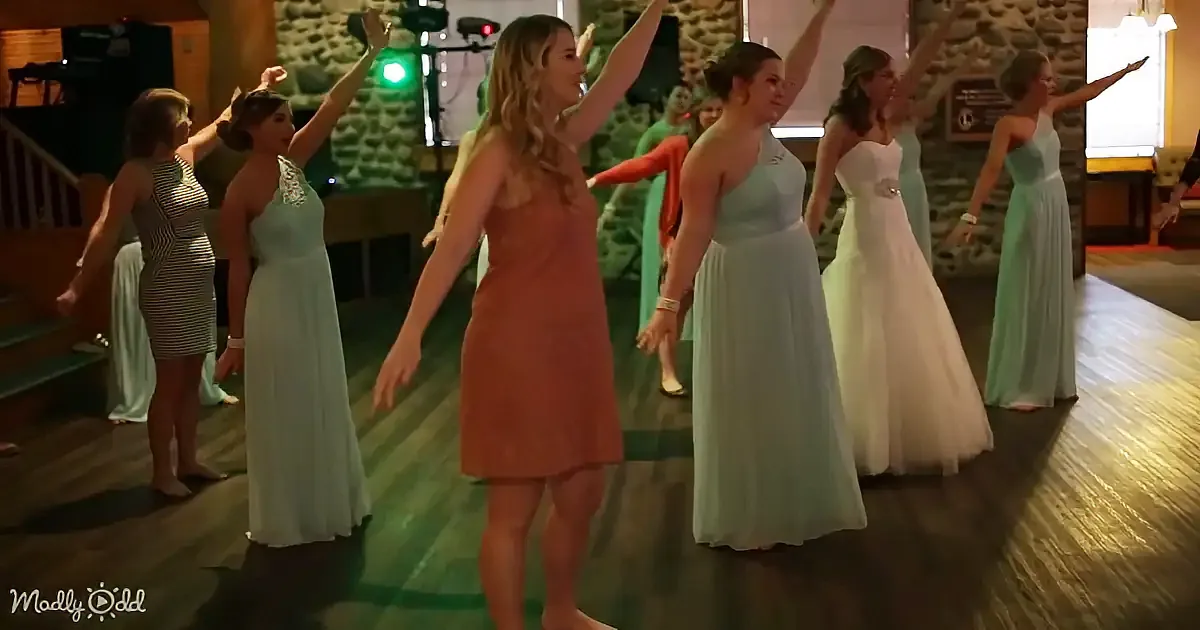 A woman in a peach dress dances on a wooden floor with others in various dresses.