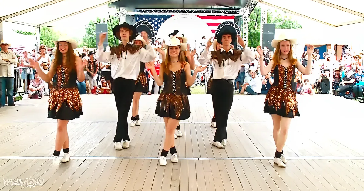 Dancers in cowboy attire perform on stage, surrounded by an audience at a festive outdoor event.