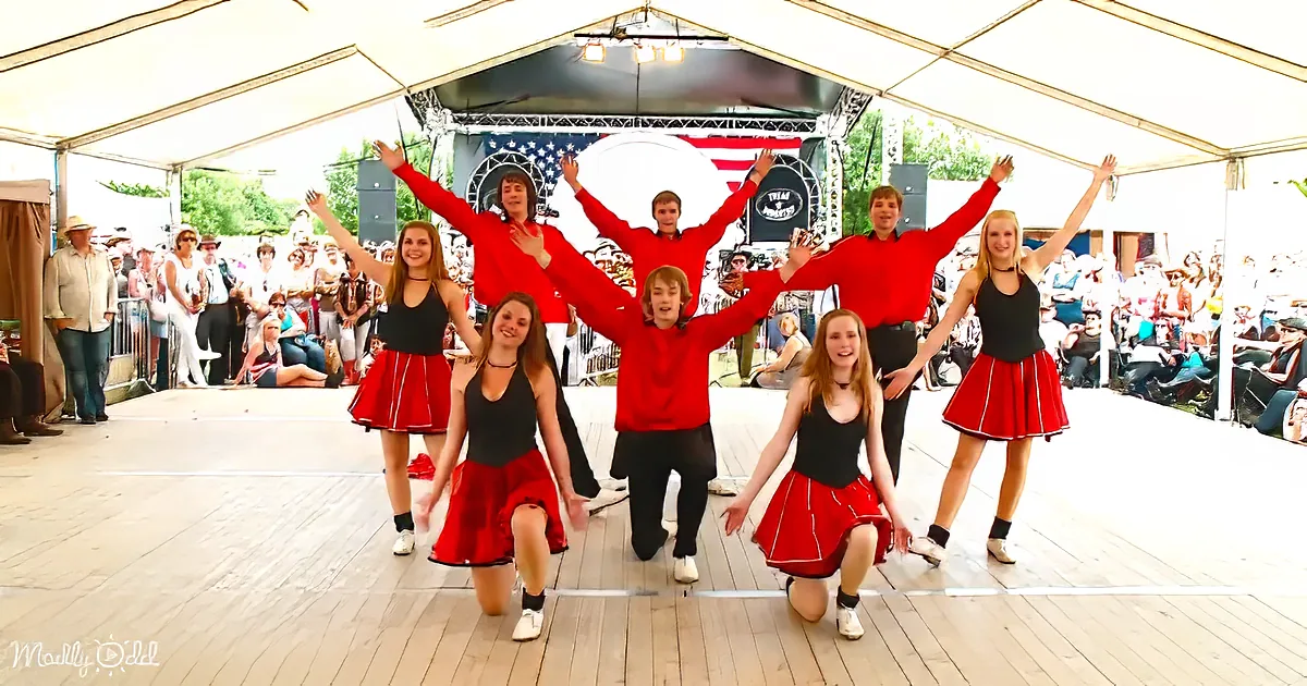 A group of dancers in red and black costumes performs on a stage in front of an American flag backdrop.