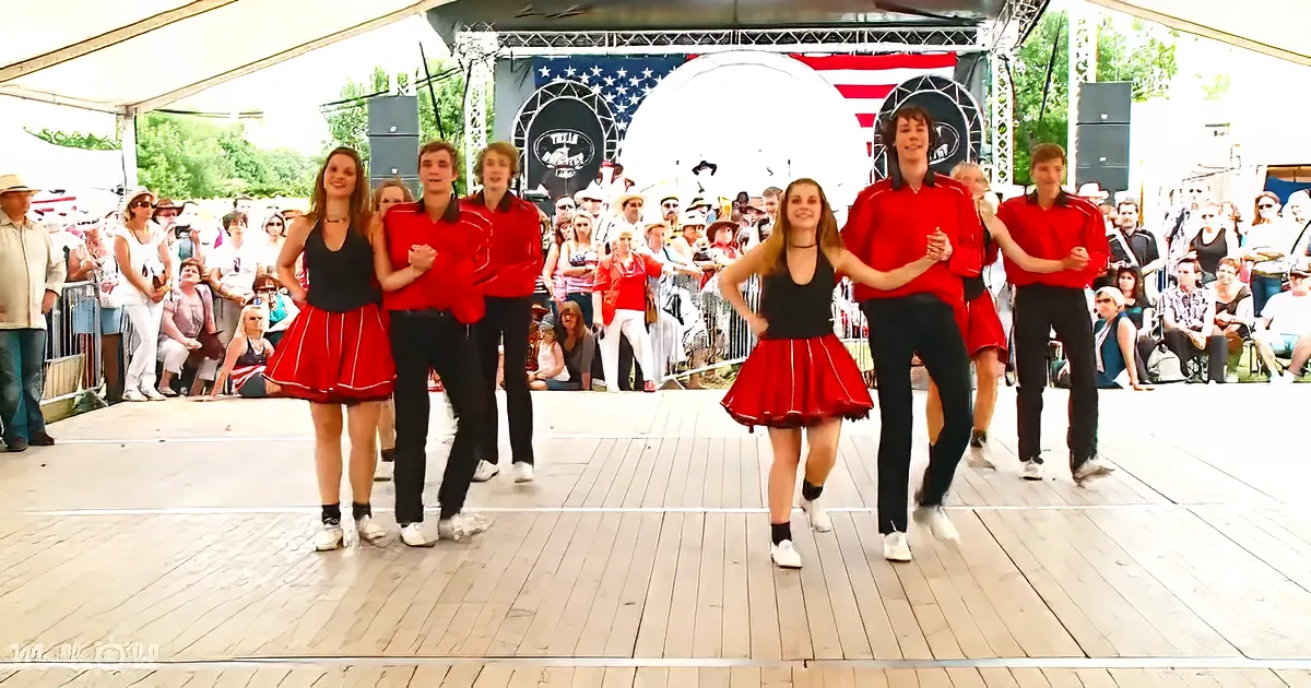 A group of dancers in red skirts and black tops perform on a stage in front of a crowd at an outdoor event.