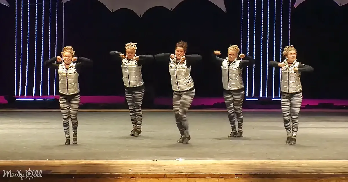 Five performers in matching silver outfits and striped pants dance on stage with synchronized arm movements.