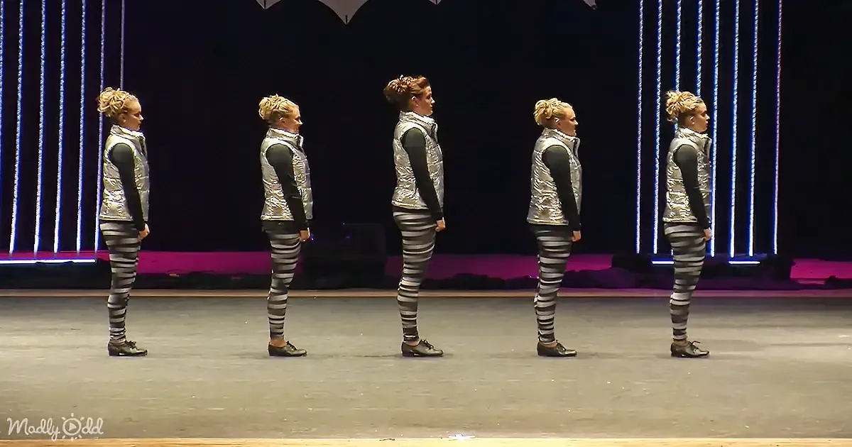 Five performers in silver vests and striped leggings stand in a row on a stage with vertical light beams behind them.