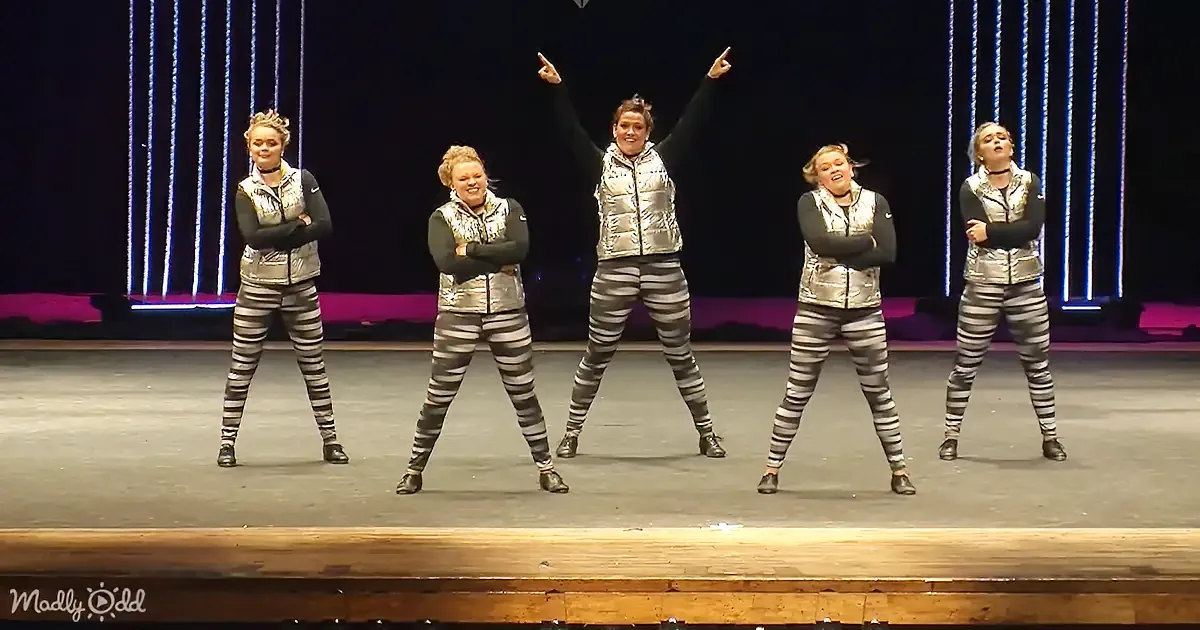 Five women in silver and black outfits perform a synchronized dance on stage with neon lights in the background.
