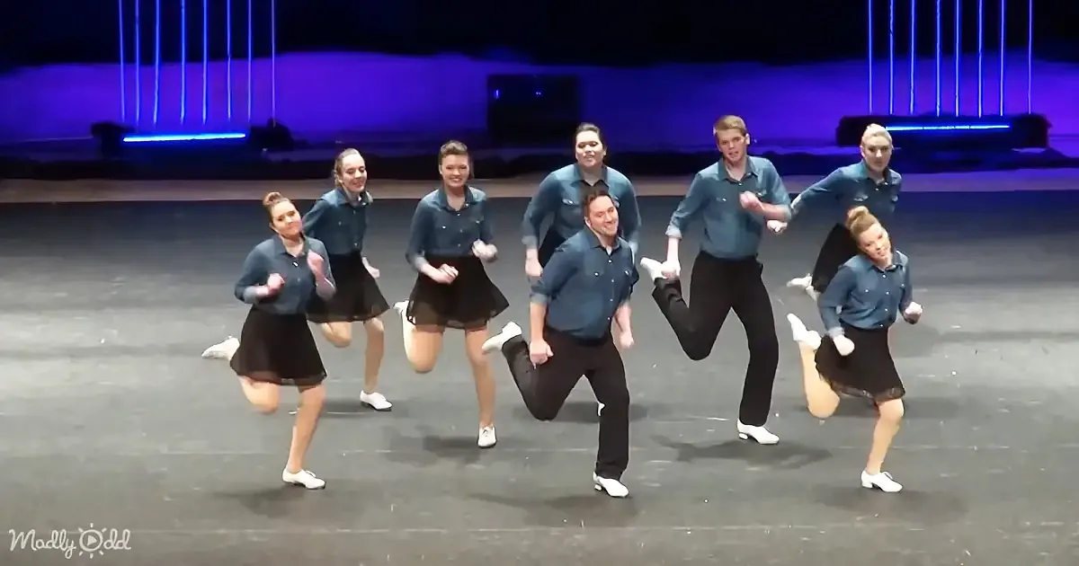 A group of dancers in blue shirts and black skirts perform a synchronized routine on stage.
