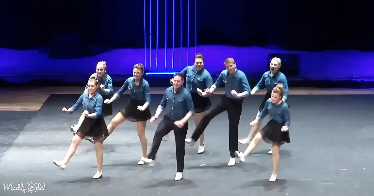 A group of dancers in blue shirts and black skirts perform a synchronized routine on stage.