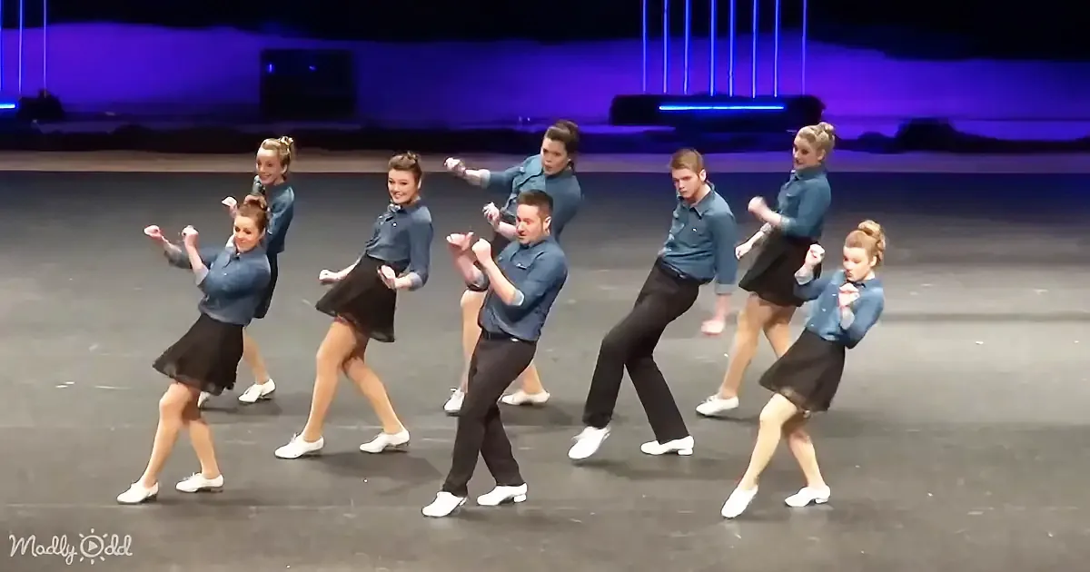 A group of dancers in blue shirts and black skirts perform a synchronized tap dance routine on stage.