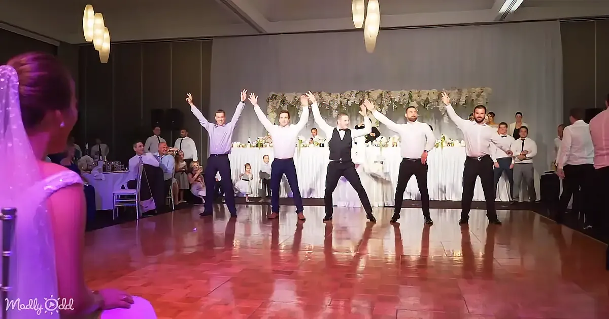 A group of men in formal attire perform a synchronized dance on a wooden floor at a wedding reception.