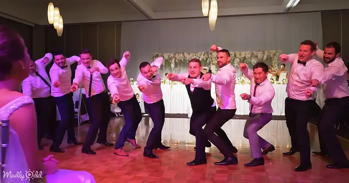 A group of men in white shirts and black pants dance energetically on a wooden floor.