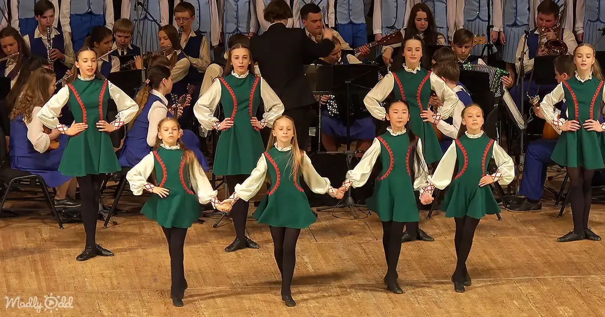 Children in green dresses perform on stage, holding hands, with a band in the background.