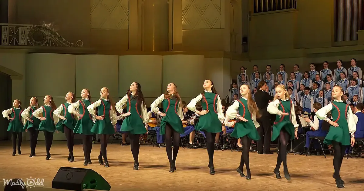 A group of dancers in green dresses perform on stage, with a choir in the background.