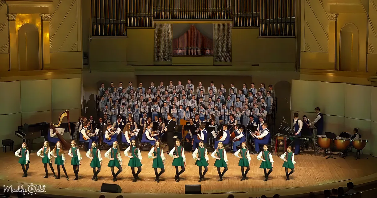A group of young dancers in green dresses perform on stage, with a large choir and orchestra behind them.
