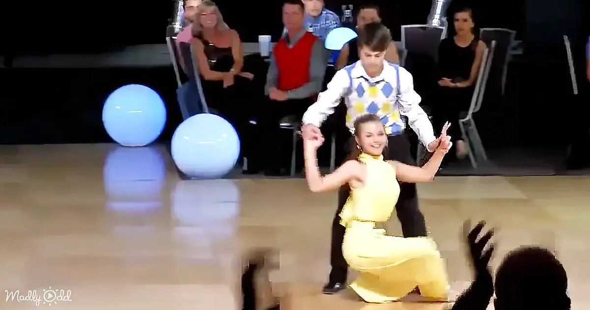 A couple in a dance pose, woman in yellow, man in white and blue, with spectators and glowing blue orbs in the background.
