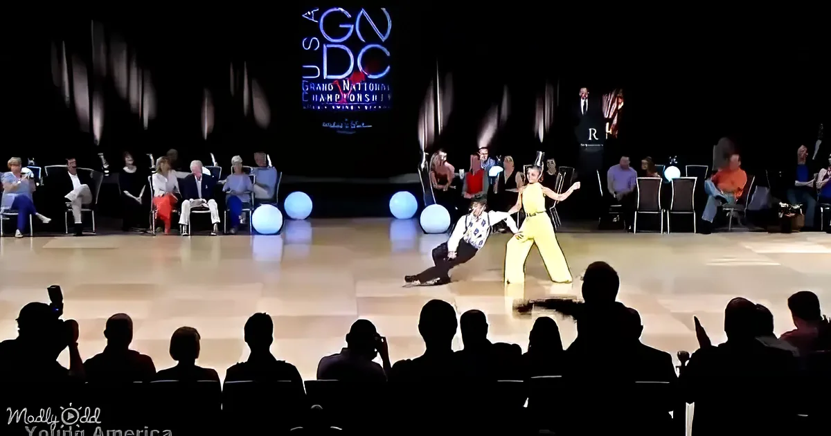 A couple in yellow and white performs a swing dance on a stage with spectators and judges in the background.
