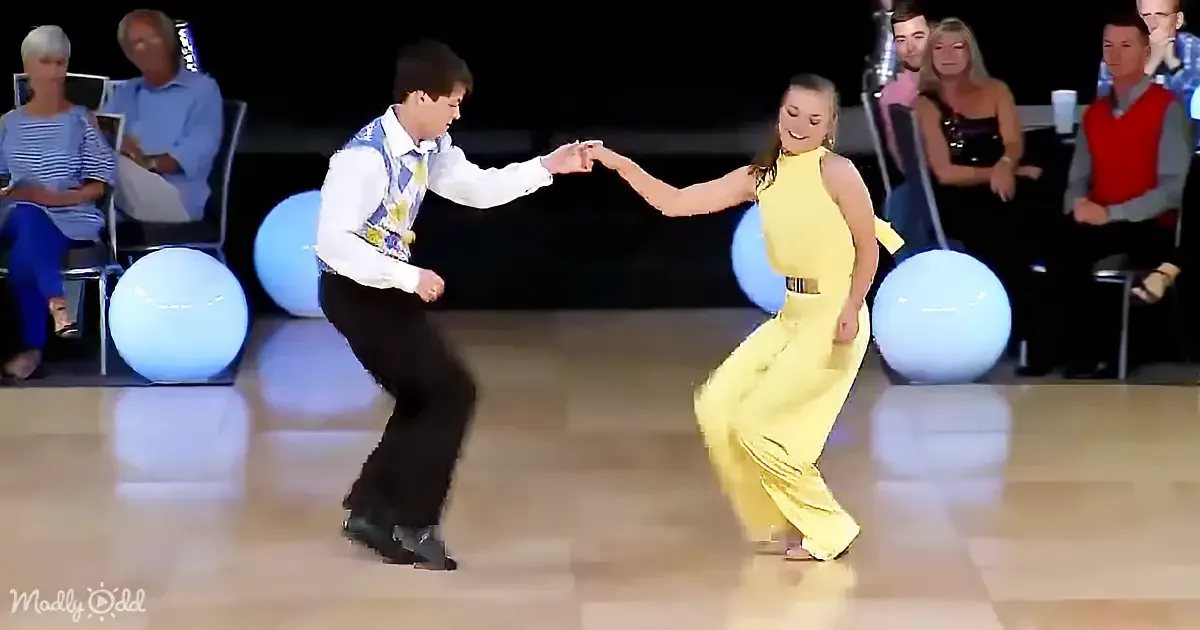 A man and woman dance gracefully on a stage, holding hands, with spectators watching in the background.