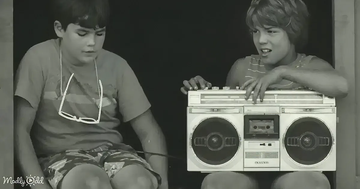 Two boys sit with a vintage boombox, one holding sunglasses, both appearing engaged.