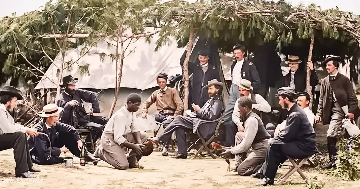 A group of men in 19th-century attire gather around a campfire, socializing under a makeshift shelter.